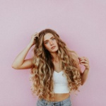 A young woman with long, wavy blonde hair, wearing a white crop top and light blue jeans, stands against a pink wall. She looks directly at the camera with a relaxed expression, her hand gently touching her voluminous hair extensions, showcasing their vibrant blonde color.