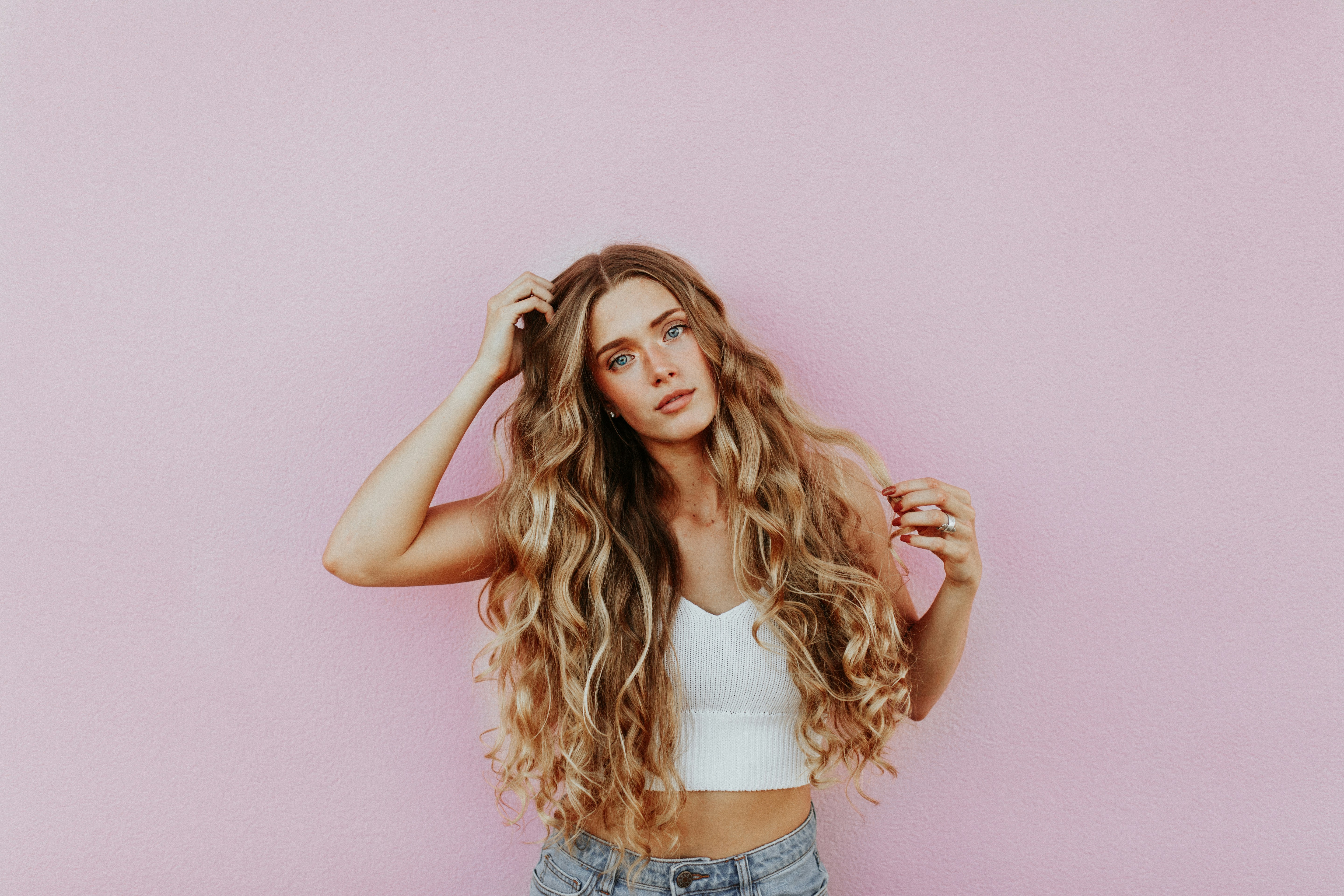 A young woman with long, wavy blonde hair, wearing a white crop top and light blue jeans, stands against a pink wall. She looks directly at the camera with a relaxed expression, her hand gently touching her voluminous hair extensions, showcasing their vibrant blonde color.