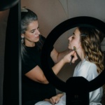 A makeup artist applies makeup to a client's face using a brush, illuminated by a ring light. The artist focuses intently while the client, seated and wearing a robe, remains still. The setting appears to be a professional studio, highlighting the expertise and attention to detail in the makeup application process. This image relates to choosing the right makeup artist in Palm Beach.