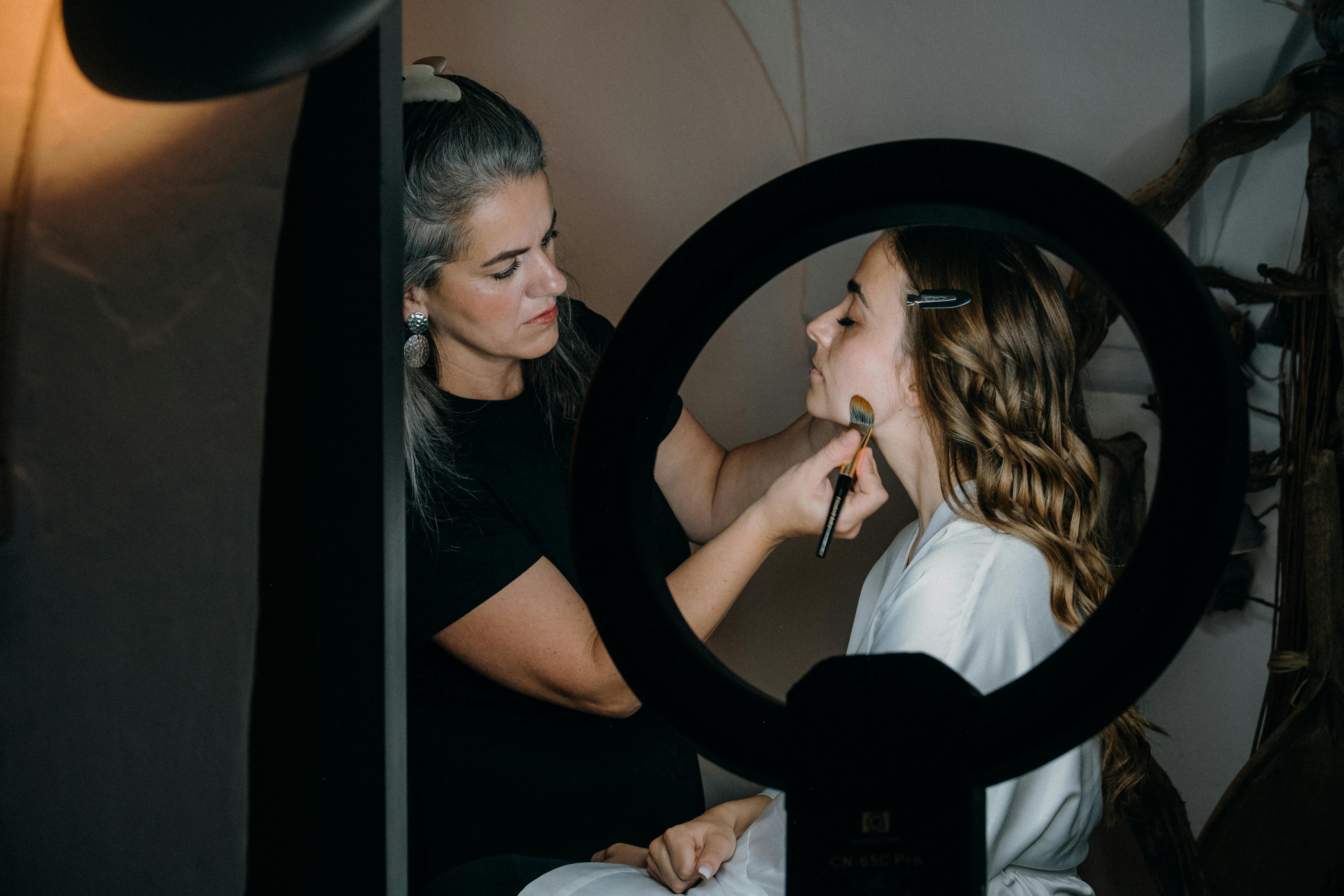 A makeup artist applies makeup to a client's face using a brush, illuminated by a ring light. The artist focuses intently while the client, seated and wearing a robe, remains still. The setting appears to be a professional studio, highlighting the expertise and attention to detail in the makeup application process. This image relates to choosing the right makeup artist in Palm Beach.