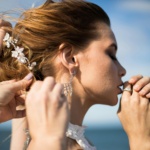 A stylist gently adjusts a bride's intricate updo adorned with floral accessories, highlighting her elegant earrings and natural makeup. The serene outdoor setting reflects the refined artistry of wedding hair and makeup offered by Palm Beach Garden salons.