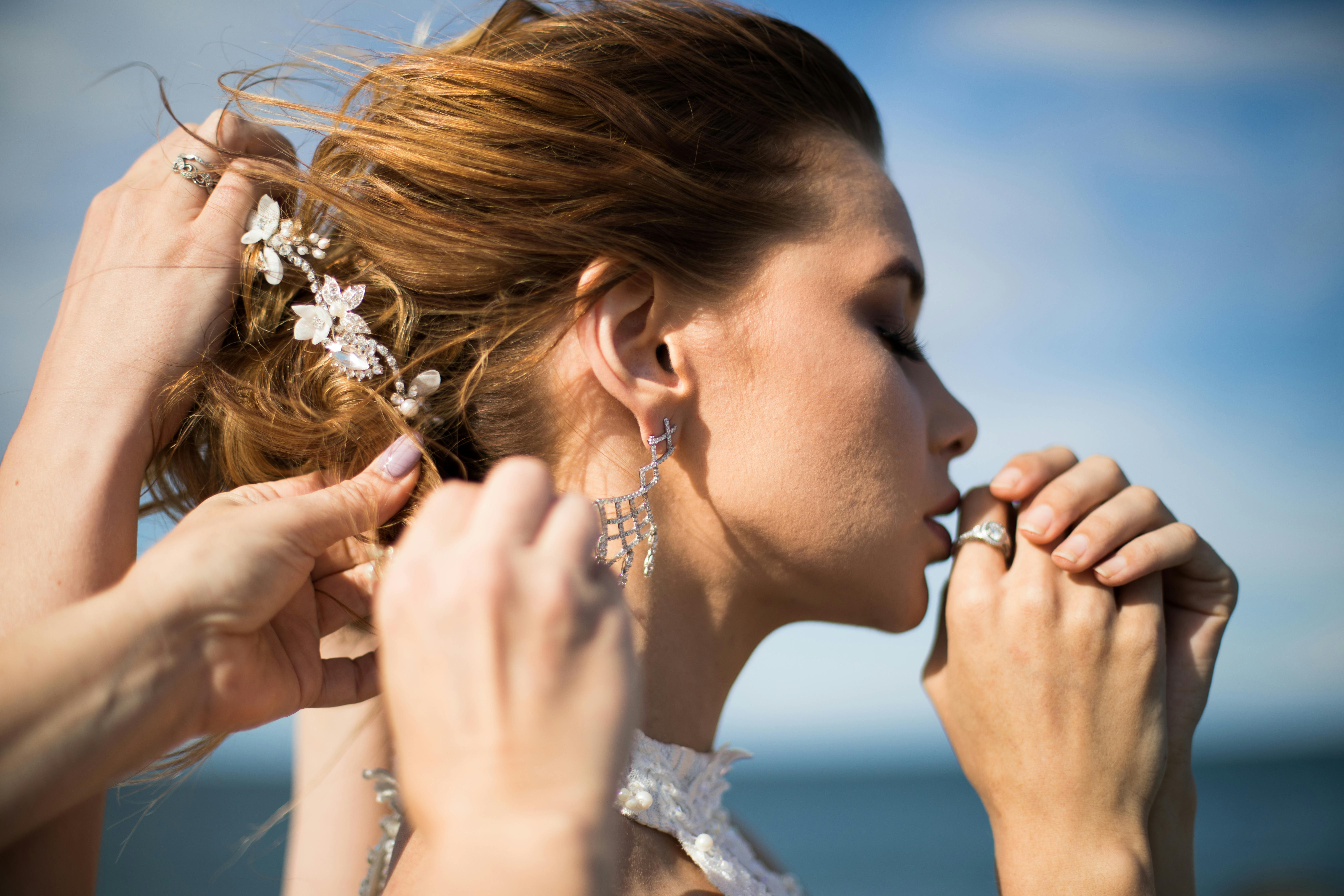 A stylist gently adjusts a bride's intricate updo adorned with floral accessories, highlighting her elegant earrings and natural makeup. The serene outdoor setting reflects the refined artistry of wedding hair and makeup offered by Palm Beach Garden salons.