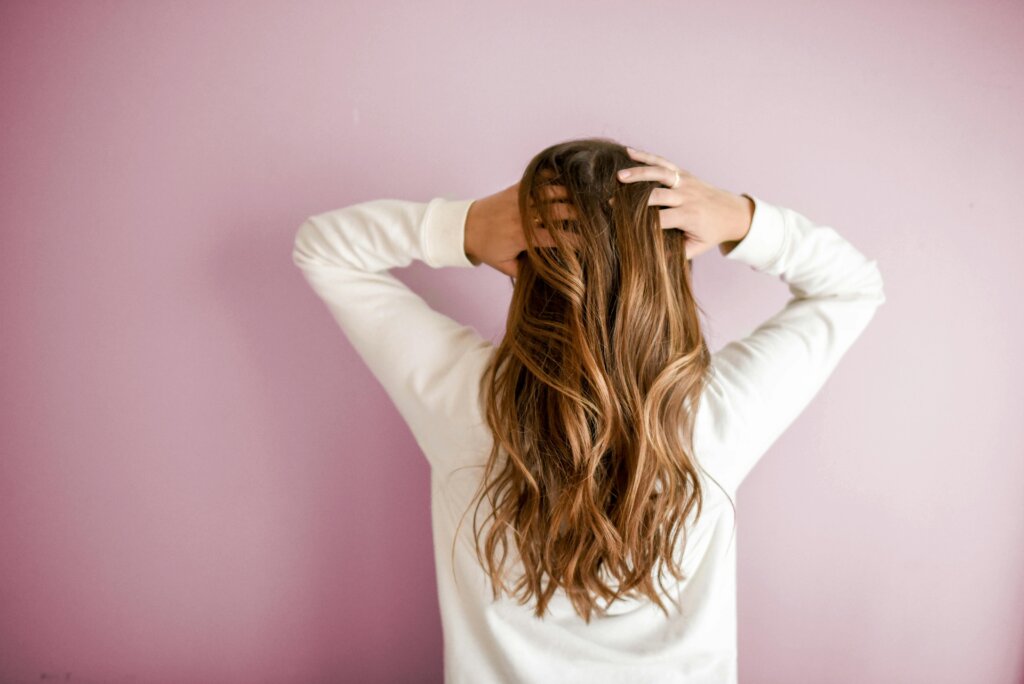 Woman with long, wavy hair wearing a white top, standing against a pink wall and running her hands through her hair, showcasing hair extensions in West Palm Beach.