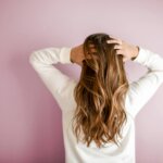 Woman with long, wavy hair wearing a white top, standing against a pink wall and running her hands through her hair, showcasing hair extensions in West Palm Beach.