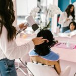 Stylist blow drying a client’s hair at a salon station, demonstrating how to prevent frizz when blow drying using a professional nozzle and sectioning technique.
