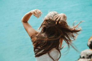 Wind-swept hair against a blue water backdrop, a reminder of the effects of summer sun and the need for hair post summer recovery.
