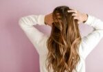 Woman with long, wavy hair wearing a white top, standing against a pink wall and running her hands through her hair, showcasing hair extensions in West Palm Beach.