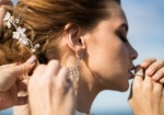 A stylist gently adjusts a bride's intricate updo adorned with floral accessories, highlighting her elegant earrings and natural makeup. The serene outdoor setting reflects the refined artistry of wedding hair and makeup offered by Palm Beach Garden salons.