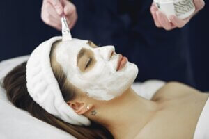 A professional aesthetician applies a white cream mask to a woman's face to determine which facial is best for glowing skin.